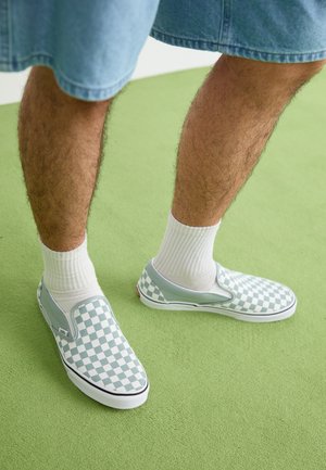 Jambes portant des chaussettes blanches et des chaussures sans lacets avec un motif à carreaux vert clair et blanc, debout sur un tapis vert.