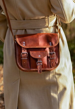 Brown leather satchel with buckles worn crossbody over a beige belt-tied coat outdoors in natural light.