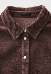 Bruine corduroy shirt met een opstaande kraag en witte drukknoopjes, gelabeld "OPUS" op het label aan de binnenkant van de kraag.