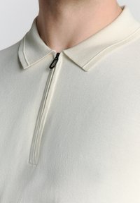 Lichtbeige polo shirt met een halve ritssluiting aan de voorkant, textuurstof en een klassiek kraagontwerp. De rits heeft een zwarte accent.