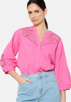 Chemisier rose à manches longues et bouffantes. Présente des broderies découpées décoratives le long des épaules et des ourlets des manches.