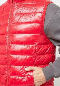 Red padded vest with a shiny finish, vertical stitch pattern, and grey sweater sleeve visible underneath. Features side pockets and a front zipper.