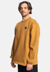 Sudadera de color mostaza hecha de una tela texturizada, con un cuello redondo acanalado y un pequeño parche negro con el logotipo en el pecho.