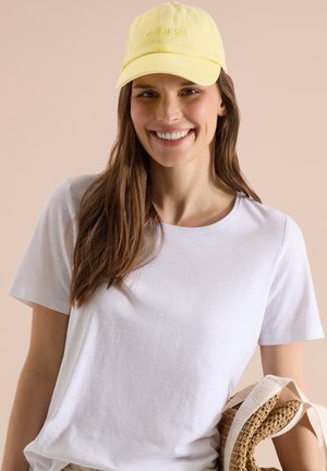 Smilende kvinde iført en gul kasket med teksten "OPTIMISME", hvid t-shirt, der holder en rullet stråhat med hvide stropper mod beige baggrund.