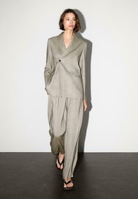 Grijs geruit pak met een oversized blazer en losse broek, gekenmerkt door een enkele zwarte knoop en lichte stof. Gedragen met zwarte slippers.