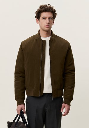 Νέος άντρας με σγουρά μαλλιά που φοράει καφέ bomber μπουφάν, λευκό πουκάμισο και μαύρο παντελόνι, κρατώντας μια μαύρη τσάντα στο αριστερό του χέρι.