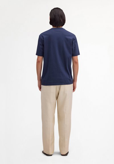 Osoba o krótkich ciemnych włosach stoi tyłem, ubrana w granatową koszulę z krótkim rękawem i beżowe spodnie na jednolitym białym tle.