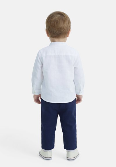 Νήπιο που στέκεται ανάσκελα, φορώντας λευκό μακρυμάνικο πουκάμισο, ναυτικό μπλε παντελόνι, λευκές κάλτσες και λευκά αθλητικά παπούτσια σε απλό φόντο.