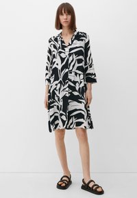 Schwarz-weißer, langärmeliger Kleid mit einem Blattmuster, Kragen und lockerer Passform, kombiniert mit schwarzen Riemchensandalen.