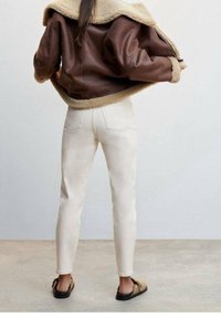 Chaqueta de cuero marrón con cuello de piel de borrego, pantalones vaqueros crema y sandalias negras. La vista trasera destaca las líneas limpias y los tonos neutros.