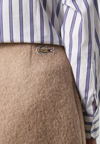 Bruine wollen rok met een zachte textuur, met daarboven gedeeltelijk zichtbaar een wit-blauw gestreept shirt, benadrukt door een geborduurd krokodillenlogo.
