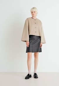 Giacca beige cropped con maniche a campana e chiusura a bottoni, abbinata a una minigonna in pelle nera e mocassini neri con suola spessa.