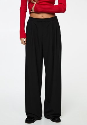 Femme portant un pantalon noir taille haute à jambes larges et un top rouge côtelé à manches longues et court, debout les bras croisés.