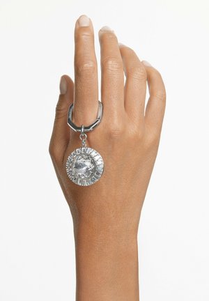 Mano con anello d'argento con grande ciondolo rotondo di gemma trasparente che pende dalla fascia su sfondo bianco.