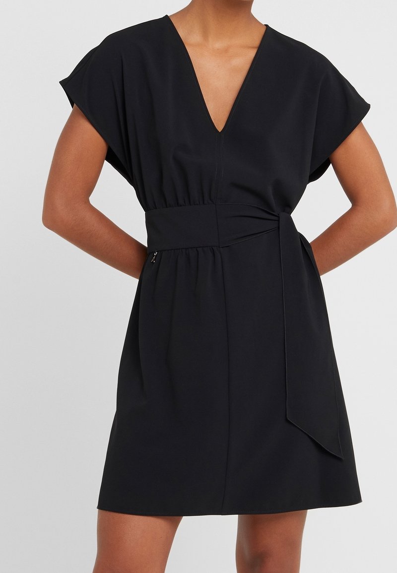 Robe noire à manches courtes avec col en V et large ceinture nouée à la taille, portée par une personne les mains posées sur les hanches devant un fond uni.