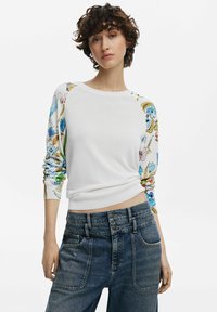 Maglione bianco con maniche a fantasia floreale, scollo rotondo e polsini a costine, abbinato a jeans in denim a vita alta con diverse tasche anteriori.