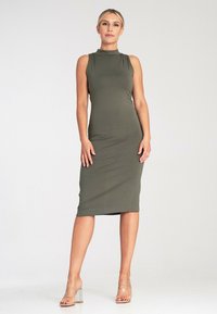 Oliwkowa, bezrękawnikowa sukienka midi w fasonie bodycon z wysokim dekoltem i gładką fakturą, w zestawieniu z przezroczystymi sandałami na słupku.