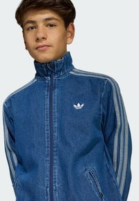 Giacca di jeans con zip in blu scuro, colletto alto, dettagli a strisce bianche e piccolo logo bianco sul petto. Due tasche laterali.