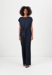 Giubbotto blu navy con maniche corte e cintura a cravatta in vita. Presenta una vestibilità comoda e un design di pantaloni ampia e fluida.