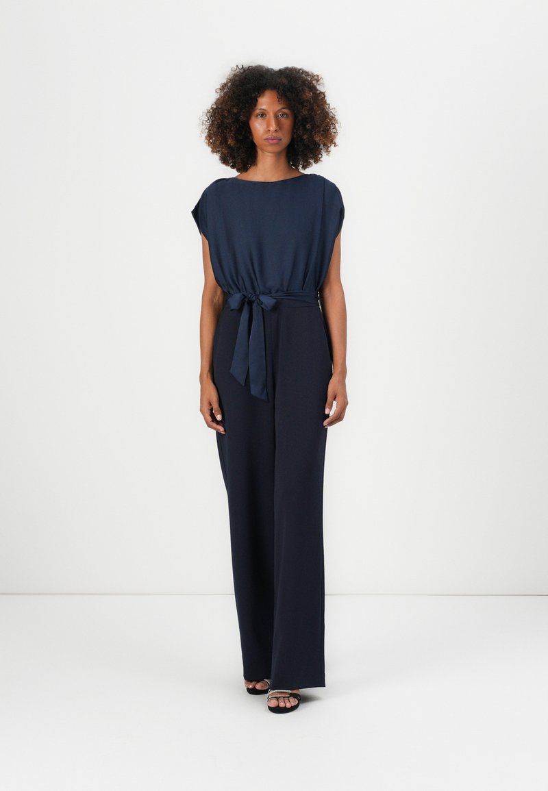 Combinaison bleu marine avec des manches courtes et une ceinture à nouer à la taille. Présente une coupe décontractée et un design de pantalon large et fluide.