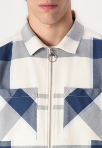Cămașă cu model în carouri, albă, bleumarin și albastru deschis. Dispune de un fermoar metalic, două buzunare aplicate și un guler punctat. Material texturat.