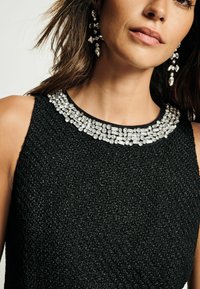 Robe noire texturée avec un col détaillé en perles. Le modèle porte des boucles d'oreilles pendantes avec des accents de gemmes transparentes.