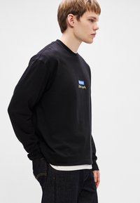 Μαύρο μπλουζάκι με μακριά μανίκια και στρογγυλή λαιμόκοψη, με μπλε και χρυσό λογότυπο. Το ύφασμα φαίνεται λείο, φορεμένο πάνω από μια λευκή μπλούζα εσωτερικά.