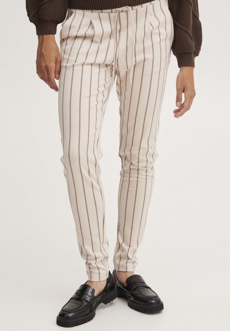 Personne portant un pantalon beige avec de fines rayures verticales foncées, un pull marron et des mocassins noirs, debout devant un fond uni.