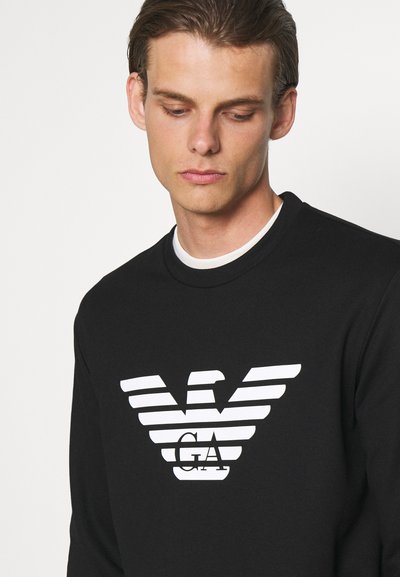 Juodas džemperis su apykakle, centre esantis didelis baltas juostuotas logotipas. Lygus paviršius ir standartinis kirpimas, paprastas dizainas.