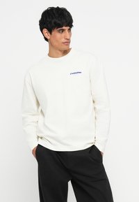 T-shirt crème à manches longues en coton avec un col rond et un logo brodé bleu sur la poitrine gauche, associé à un pantalon noir.