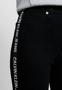 Svarta denimjeans med en sidpanel som har "CALVIN KLEIN JEANS"-logotyp i vitt. Slim fit med standardfickor och knappstängning.