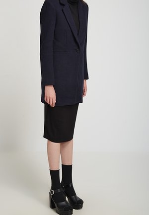 Frau steht und trägt einen schwarzen knielangen Rock, einen schwarzen Rollkragenpullover, einen dunkelblauen, geknöpften Blazer, schwarze Knöchelsocken und schwarze High Heels.