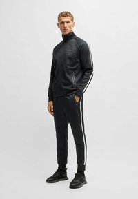 Svart idrottsdräkt med en zip-up jacka med hög krage, texturerat tyg och vita sidostråkar; parat med matchande byxor.