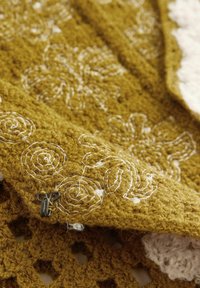 Il tessuto lavorato a maglia color senape presenta ricami floreali bianchi, un filato testurizzato e un dettaglio con chiusura metallica decorativa. Il motivo a punto aperto è predominante.