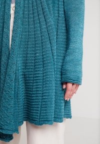 Teal stickad cardigan med ribbad textur och spetsdetaljer vid fållen. Har långa ärmar och en avslappnad passform, bärs över lätt tyg.