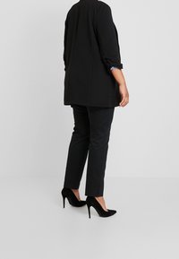 Czarna, oversize'owa marynarka, czarne dopasowane spodnie oraz czarne zamszowe wysokie obcasy. Marynarka ma podwinięte rękawy i prosty fason, na jednolitym tle.