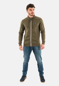Felpa verde oliva con zip, colletto alto, cordini regolabili, tasche frontali, zip in metallo ed emblema sul lato sinistro del petto.