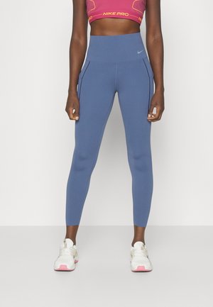Legging taille haute bleus avec poches latérales, fabriqués dans un matériau extensible. Associés à des baskets blanches avec des accents roses.