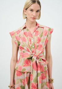 Blusa a stampa floreale nei toni di rosa, verde e bianco. Maniche corte, chiusura con bottoni sul davanti e dettaglio a vita annodata. Tessuto leggero con una finitura testurizzata.