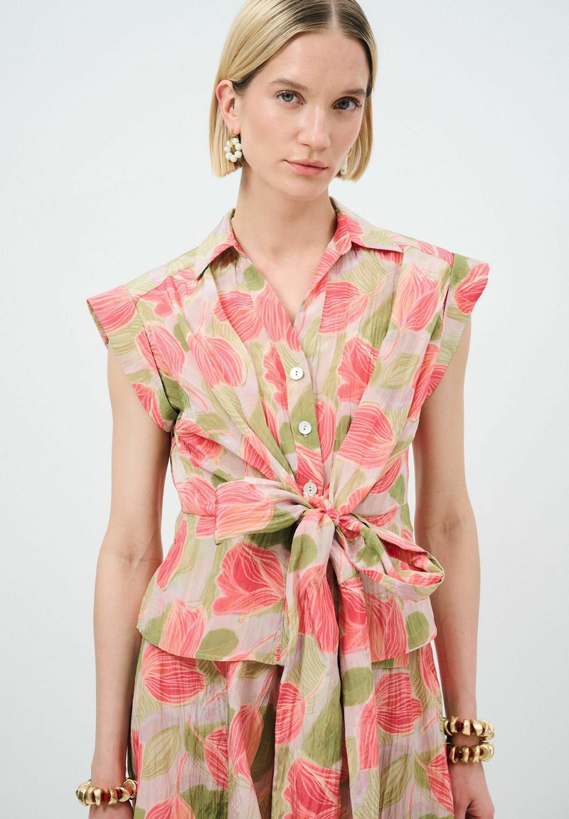 Bloes met bloemenprint in roze, groen en wit. Korte mouwen, knoopsluiting aan de voorkant en een strikdetail in de taille. Lichtgewicht stof met een gestructureerde afwerking.