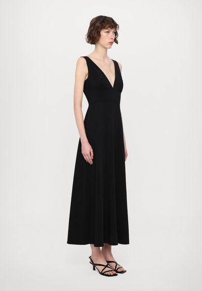 Lang zwarte jurk met een diepe V-hals, aansluitende bodice en zwierige rok. Gecombineerd met zwarte sandalen met banden. Soepele stoftextuur.