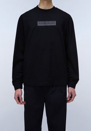 Personne portant un t-shirt à manches longues noir avec un logo rectangulaire gris indiquant Napapijri Geographic sur la poitrine et un pantalon noir.