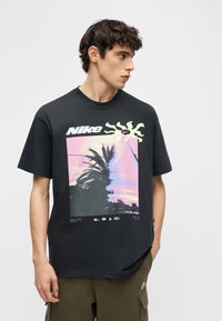 Νέος άνδρας που φοράει μαύρο T-shirt της Nike με γραφικό αποτύπωμα φοίνικα και ήλιου σε παστέλ χρώματα, στέκεται μπροστά σε απλό φωτεινό γκρι φόντο.