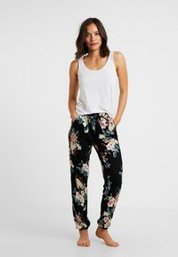 Zwarte stoffen pyjamabroek met kleurrijke bloemenpatronen, een elastische tailleband met trekkoord en een relaxte pasvorm. Gekleed met een wit hemdtopje.
