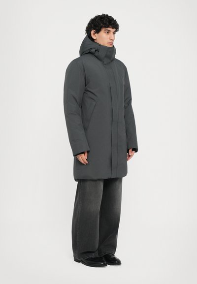 Pelēks izolēts parka ar augstu apkakli un kapuci, ar plūstošu dizainu, lielām kabatām, valkāts kopā ar platām melnām jeans un apaviem.