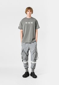 Camiseta gris con letras "MWM", combinada con pantalones cargo grises que presentan cremalleras y acentos. Unos zapatos de tela negros con textura y sin cordones completan el conjunto.
