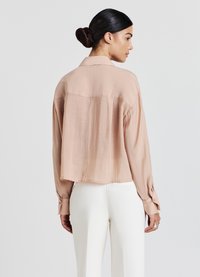 Blusa leggera di colore rosa pallido, a maniche lunghe e dal taglio corto, con una texture sottile e polsini abbottonati, abbinata a pantaloni bianchi aderenti.
