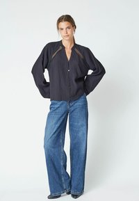 Blusa nera strutturata con scollatura a V e chiusura a bottone, con dettagli in rete sulle spalle. Abbinata a jeans in denim blu a gamba larga.