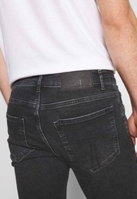 Mörka jeans i denim med rak passform, fem fickor och en texturerad läderlapp vid midjan. Diskret blekning och sömnadsdetaljer synliga.