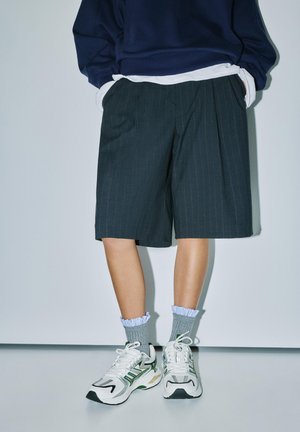 Personne portant un short sombre à fines rayures jusqu’aux genoux, des chaussettes grises à volants, des baskets blanches et vertes, un sweat-shirt bleu marine et des couches de chemise blanche.